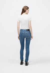 Gai krāsas pieguļošs T-krekls, kopā ar tumši ziliem šaurajiem džinsiem, uzsverot gludu audumu ar smalku šuvumu un aizmugurējo jostasvietas logotipu.