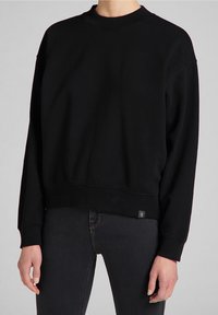 Persona vestida con un sudadera negra de manga larga lisa y jeans oscuros ajustados, de pie contra un fondo gris neutro.