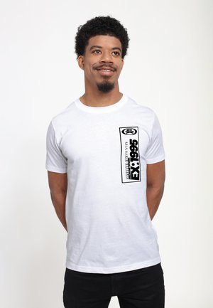 Hvid bomulds t-shirt med korte ærmer, der har et stort sort grafisk print på fronten og et lille logo i øverste venstre hjørne.