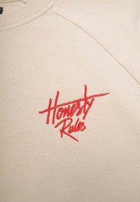 Beige sweatshirt med teksten "Honesty Rules" i store broderede bogstaver i rød, med ribkant i halsen og tekstureret stof.