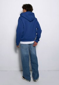 Niebieska bluza z kapturem, wykonana z miękkiego materiału, charakteryzująca się luźnym krojem z prążkowanymi ściągaczami i dołem, w zestawieniu z jasnoniebieskimi dżinsami.