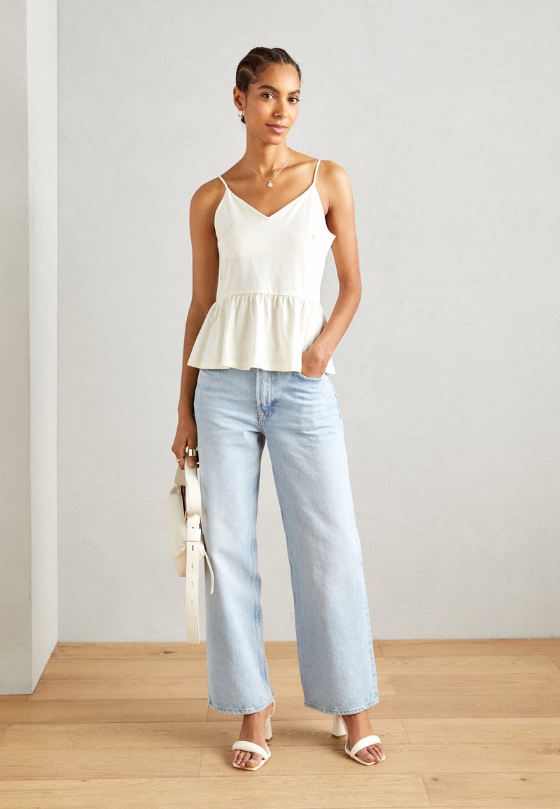 Top peplum blanco con tirantes finos, combinado con pantalones vaqueros de pierna ancha de color azul claro. La modelo sostiene un bolso blanco y lleva sandalias blancas.
