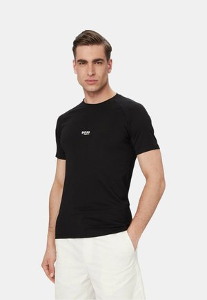 Uomo che indossa una t-shirt nera BOSS a maniche corte e pantaloncini bianchi, in piedi con una mano in tasca su sfondo semplice.