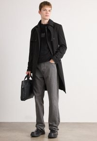 Black trench coat over a black T-shirt, gray jeans, and black boots; holding a black bag. Smooth fabric, tailored design, and neutral colors.