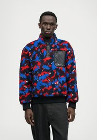 Fleece pullover in red, blue, and black camo pattern with half-zip, elastic cuffs, and a zippered black chest pocket featuring a logo.