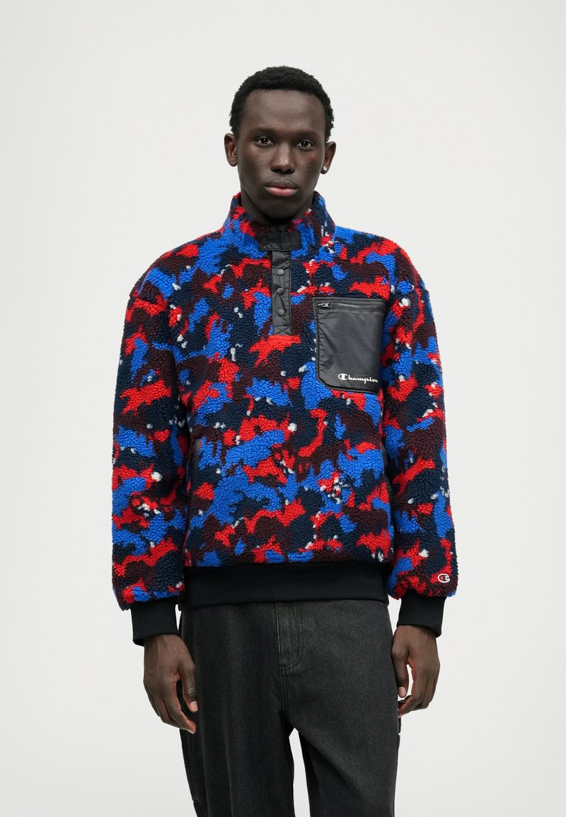 Fleece pullover in red, blue, and black camo pattern with half-zip, elastic cuffs, and a zippered black chest pocket featuring a logo.