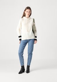 Krémovo-biela mikina s kapucňou a rebrovaným lemom, doplnená čiernymi pruhmi, spárovaná s modrými džínami a čiernymi topánkami na podpätku, drží zelenú tašku.