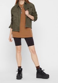Frau, die eine olivgrüne Jacke, einen braunen langen Pullover, schwarze Biker-Shorts, braune Socken und schwarze Plateaustiefel mit Schnürung trägt, steht vor einem weißen Hintergrund.