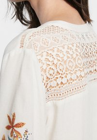 Närbild av en beige blusaxel med intrikata spetsdetaljer och ett blommönster på ärmen, bärs av en person med kort brunt hår.