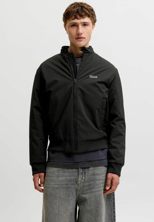 Jeune homme portant une veste noire zippée, un t-shirt foncé en dessous et un jean gris clair, debout devant un fond clair uni.