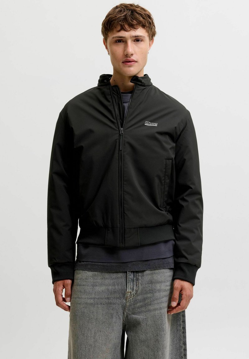 Jeune homme portant une veste noire zippée, un t-shirt foncé en dessous et un jean gris clair, debout devant un fond clair uni.