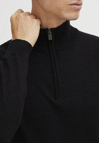 Svart strikket genser med halv-glidelås. Har ribbede mansjetter og krage, glatt tekstur, og en slank, figursydd design.