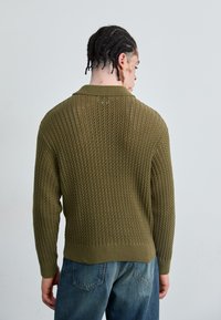 Olive green knitted sweater with a textured design, featuring a ribbed collar and cuffs, and open weave patterns along the body.