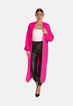 Lange cardigan met open voorkant in felroze breiwerk, met ribbelstructuur. Gepaard met zwarte leren joggers en een witte top. Puntige zwarte hakken.