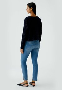 Marineblaue Kabelstrickpullover, verkürztes Design; mittelhohe blaue Skinny Jeans; schwarze Slingback-Schuhe mit spitzer Zehe und niedrigem Absatz; neutraler Hintergrund.