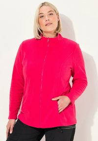 Žena s krátkými blond vlasy, oblečená v jasně růžovém fleecovém saku s upnutým zipem, s jednou rukou v kapse saka, stojící na jednoduchém pozadí.