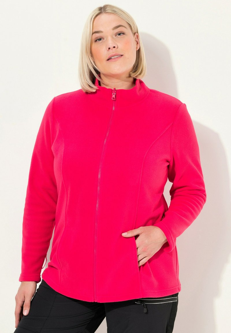 Žena s krátkými blond vlasy, oblečená v jasně růžovém fleecovém saku s upnutým zipem, s jednou rukou v kapse saka, stojící na jednoduchém pozadí.