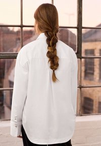 Hvid langærmet skjorte med krave, der har en rygfald, knapmanschetter og en afslappet pasform, båret over mørke bukser.