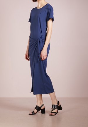 Femme portant une robe midi bleue à manches courtes avec un détail de nœud sur le côté, associée à des sandales noires à talons blocs, debout sur un sol beige.