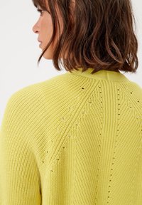 Žlutý pletený svetr s texturovaným vzorem a šikmými liniemi, s ventilačními otvory na zadní straně. Hladký límec a manžety.