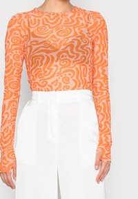 Lange mouwen bovenstuk van doorschijnende oranje stof met een wervelpatroon. Gecombineerd met witte, taillebroeken met een aansluitend ontwerp.