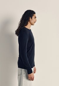 Čierna priliehavá košeľa s dlhým rukávom, s jemnou textúrou a okrúhlym výstrihom. Nosená s bledými denimovými nohavicami, viditeľná z boku.