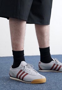 Grå sportsko med maroon striber, gummisål og snørebåndsdesign, parret med sorte strømper og knælange sorte shorts på et blåt tæppe.