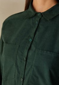 Camicia verde scuro a maniche lunghe con apertura a bottoni, colletto, due tasche sul petto e tessuto strutturato con bottoni coordinati.