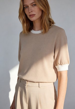 Jeune femme aux cheveux châtain clair portant un pull beige à manches courtes avec des poignets blancs et un pantalon beige taille haute, debout devant un fond uni.
