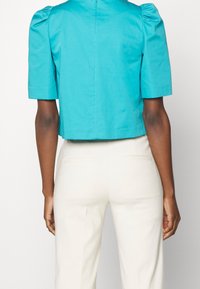 Vue de dos d'une personne portant un chemisier turquoise à manches courtes avec légèrement épaules bouffantes et un pantalon blanc taille haute avec poches arrière.