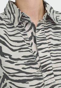 Chemise col en tissu gris clair avec un motif rayé noir de zèbre. Coutures visibles et design de patte de boutonnage au niveau du cou.