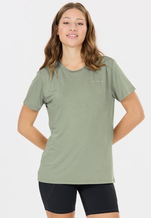 Junge Frau mit gewelltem Haar steht mit den Händen hinter dem Rücken, trägt ein olivgrünes T-Shirt und schwarze Radhosen vor einem schlichten weißen Hintergrund.