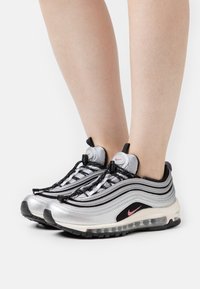Stříbrné a černé tenisky Nike Air Max 97 s elegantním designem, texturovaným svrškem a viditelným vzduchovým odpružením v podešvi. Součástí jsou elastické tkaničky.