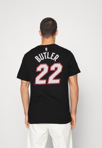 Mężczyzna w czarnej koszulce koszykarskiej z napisem "BUTLER" i numerem "22" w kolorze czerwonym i białym, stojący na tle jednolitego białego koloru.