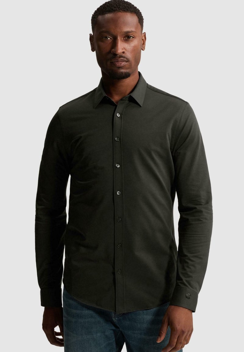 Homme portant une chemise boutonnée à manches longues vert foncé et un jean bleu, debout devant un fond clair uni.