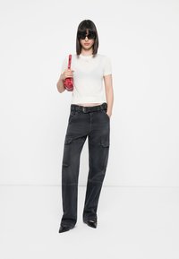 Gekürztes cremefarbenes Strickoberteil, schwarze Cargo-Hosen mit Taschen, schwarzer Gürtel, rote Handtasche mit Muster und schwarze spitze Absätze.