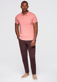 Růžová polo košile s kapsou, spárovaná s hnědými kalhotami a světle béžovými slip-on botami. Hladká tkanina, klasický střih, minimalistický design.