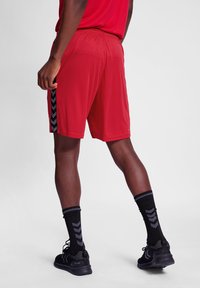 Pantalones cortos deportivos rojos hechos de material liviano, con un patrón de chevrón negro a lo largo de los costados, combinados con calcetas y calzado negros.