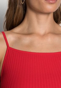 Červený žebrovaný top bez rukávů s tenkými ramínky, který má čtvercový výstřih. Hladká textura zdůrazňuje elasticitu látky. Jsou viditelné zlaté kruhové náušnice.