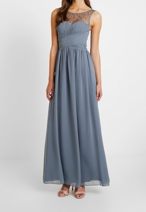 Mujer vistiendo un vestido largo sin mangas azul grisáceo con escote transparente con abalorios y corsé fruncido ajustado, posando frente a un fondo sencillo.