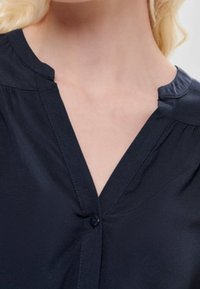 Blouse bleu marine en tissu lisse, présentant un col en V et de subtils plis, avec une fermeture à bouton unique au niveau de l'encolure.