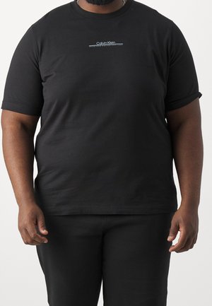 Muž nosící černé tričko Calvin Klein a černé kalhoty, stojící na jednobarevném světlém pozadí.