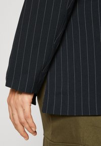 Námořnický blazer s jemným pruhovaným vzorem, vyrobený z lehce strukturované látky, s bílými pruhy a bočním rozparkem na spodním lemu; ruka viditelná na rukávu.