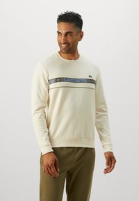 Mann iført kremfarget sweatshirt med blå og olivenstripe og olivenbukser, smilende og ser mot venstre mot hvit bakgrunn.