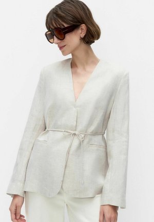 Nainen, joka pukeutuu ylisuuriin ruskeisiin aurinkolaseihin ja vaalean beigeen pellavapaitaan, jossa on ohut vyö sidottuna vyötärölle.