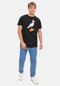 Black cotton t-shirt featuring a graphic of a white bird with an orange accent, paired with light blue jeans and white sneakers.