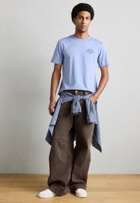 Ανοιχτό μπλε βαμβακερό μπλουζάκι με λογότυπο, καφέ παντελόνι φαρδιάς γραμμής και μπλε καρό πουκάμισο δεμένο στη μέση· λευκά αθλητικά παπούτσια πάνω σε ξύλινο πάτωμα.