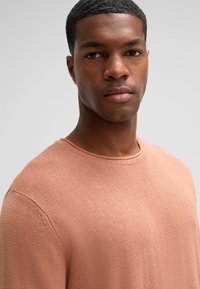 Maglione rosa chiaro con un tessuto a maglia testurizzato, scollo a girocollo e vestibilità rilassata. Presenta maniche corte e una cucitura visibile sulla spalla.