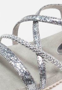 Sandali glitter argento con cinturini incrociati, dettagli intrecciati e una soletta in suede morbido. La superficie testurizzata aggiunge interesse visivo.