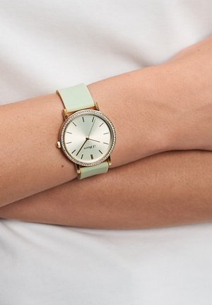 Gold-tone wristwatch with a mint green silicone strap, round face, and a bezel with clear crystals; features minimalistic hour markers.